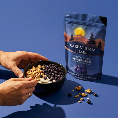 Hands holding a bowl of granola with freeze-dried whole blueberries next to pouch on a blue background