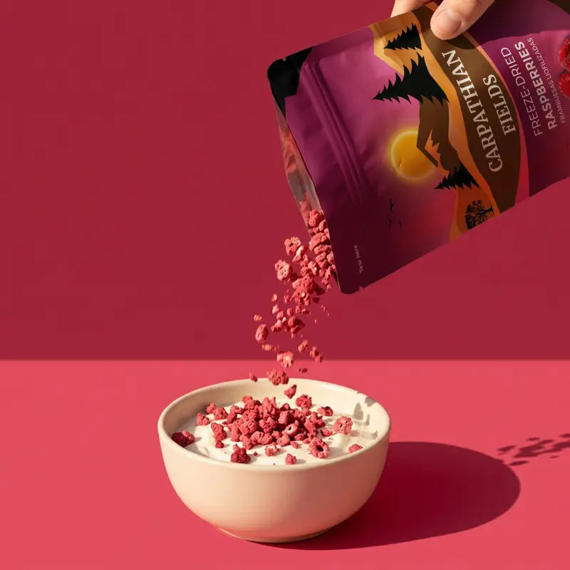 Hand pouring Carpathian Fields freeze-dried raspberry crumbs into a bowl of yogurt on a pink background